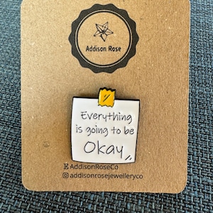 May include: White enamel pin with a yellow sticky note that reads "Everything is going to be okay." The pin is on a brown card with the text "Addison Rose" in a black circle.