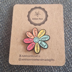 Puede incluir: Un pin de margarita colorido con palabras positivas como "amado", "valioso" y "único" en los pétalos. El pin está sobre una tarjeta marrón con el texto "Addison Rose" y detalles de redes sociales.