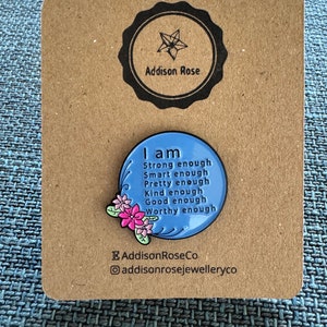 以下が含まれることがあります： 白の縁取りとピンクの花が付いた青いエナメルピン。ピンには「私は十分に強い 十分に賢い 十分に美しい 十分に親切 十分に良い 十分に価値がある」と書かれています。