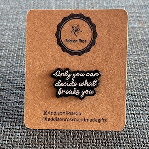 Puede incluir: Un pin de esmalte negro con el texto "Only you can decide what breaks you" en escritura blanca. El pin está sobre una tarjeta marrón con un logotipo circular negro y el texto "Addison Rose". La tarjeta también tiene el texto "AddisonRoseCo" y un identificador de Instagram.