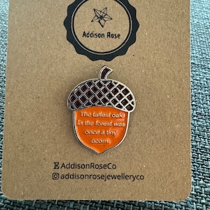 Puede incluir: Un pin de bellota de plata con fondo de esmalte naranja. El pin dice "El roble más alto del bosque fue una vez una pequeña bellota." El pin está en una tarjeta marrón con el texto "Addison Rose" y "AddisonRoseCo addisonrosejewelleryco".