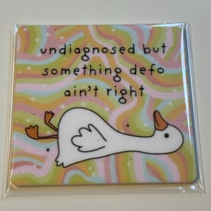 Op de afbeelding: Vierkante onderzetter met een witte ganzenillustratie en de tekst "undiagnosed but something defo ain't right." De achtergrond heeft golvende strepen in pastelkleuren. De onderzetter zit in een doorzichtige plastic hoes.