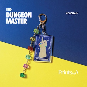 Può includere: Un portachiavi rettangolare in acrilico con un design a carta blu, un'illustrazione di una mano bianca e il testo THE DUNGEON MASTER. È fissata una catenella dorata, decorata con sei perline cubiche traslucide multicolori in fila.