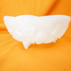 May include: A white milk glass bowl with a floral design and three legs. The bowl is on a yellow surface.