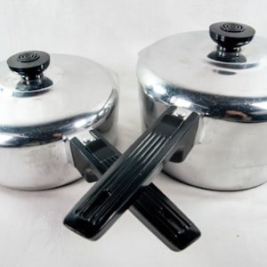 May include: Two vintage aluminum pots with black handles and lids. The lids have black knobs. The pots are sitting on a white surface. Kitchen cookware.