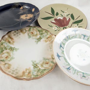May include: Four vintage ceramic plates with floral designs. The plates are in various colors, including blue, green, and white. The plates have gold trim and are in good condition.