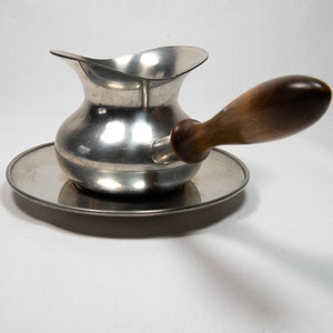 May include: A silver pitcher with a wooden handle and a matching silver saucer. The pitcher has a rounded body and a wide, flared lip.