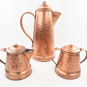 May include: A set of three copper kitchenware items: a tall hammered-finish coffee pot with a lid and handle, and two smaller matching pots with lids and handles. The copper has a warm, reddish-brown hue.