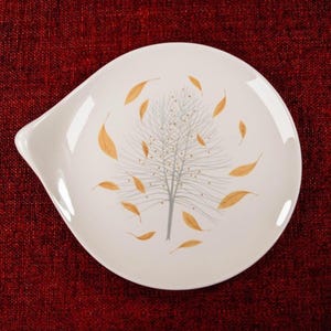 May include: A white ceramic spoon rest with a tree and falling leaf design. The tree is gray with gold accents, and the leaves are gold. The spoon rest has a curved handle on one side.