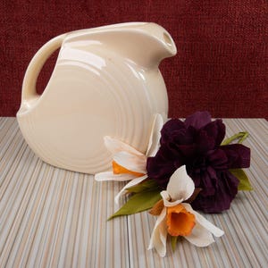 May include: A cream-colored ceramic pitcher with a curved handle and a spout, featuring concentric ring detailing on its side. Beside the pitcher are two white and orange daffodils and a large, dark purple paper flower with green leaves.