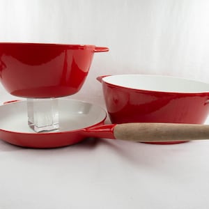 May include: A set of three red enameled cookware pieces. The set includes two bowls and a frying pan with a wooden handle. The interiors are white. The cookware is displayed on a white background.
