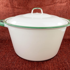 May include: White enamelware pot with a lid and green trim. The pot has two handles and a handle on the lid. The pot is sitting on a red-brown surface. The pot is a classic kitchenware item.
