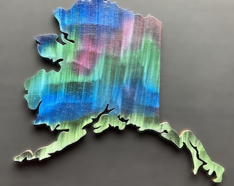 May include: Wooden cutout of the state of Alaska, painted with a vibrant depiction of the Northern Lights. The artwork features shades of blue, green, and pink, creating a colorful and eye-catching design. The state outline is approximately 30 cm in length.