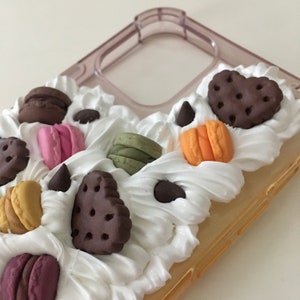 May include: A clear phone case decorated with white frosting, chocolate chip cookies, macarons, and other sweet treats.
