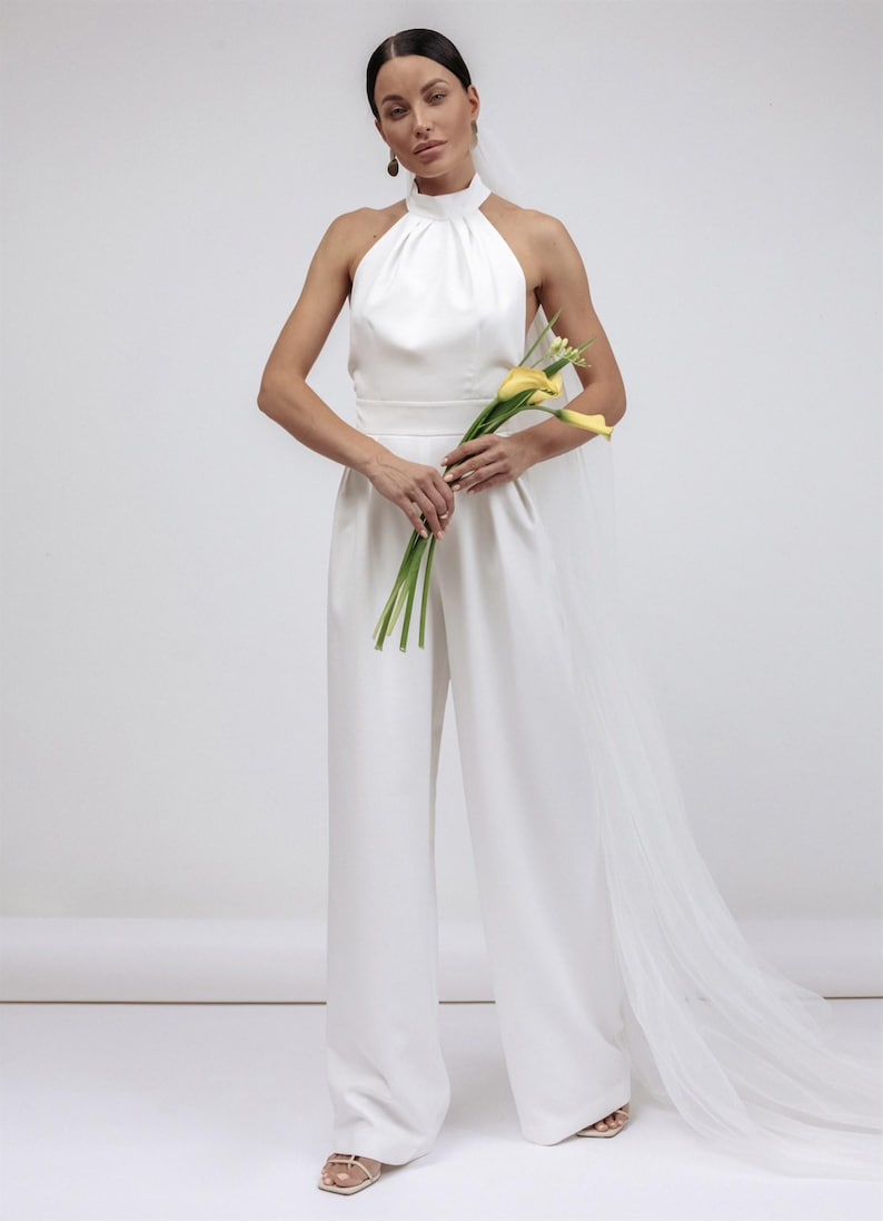 May include: A white bridal jumpsuit with a halter neckline and wide-leg pants. The outfit includes a sheer veil and is accessorized with yellow calla lilies. The overall style is minimalist and elegant.