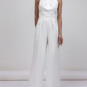 May include: A white jumpsuit featuring a halter neckline and wide-leg pants. The top has a high, gathered collar and a fitted waist. The pants are floor-length. The outfit is made of a smooth, flowing fabric, creating a sleek, elegant silhouette.