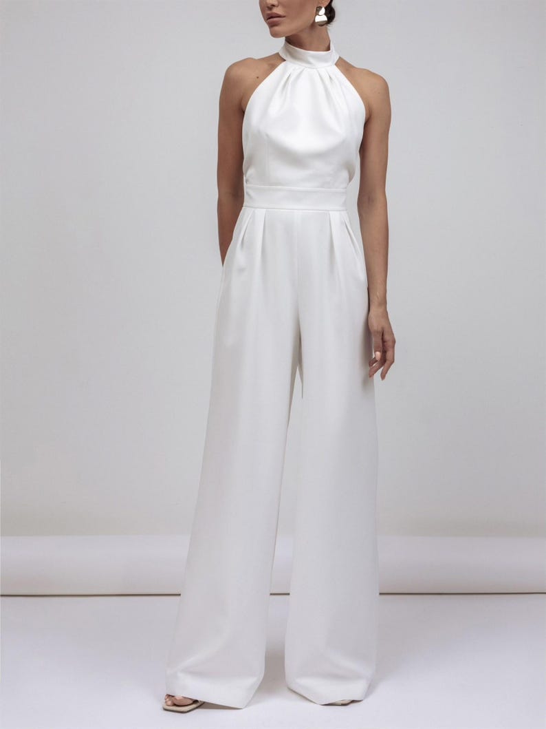 May include: A white jumpsuit featuring a halter neckline and wide-leg pants. The top has a high, gathered collar and a fitted waist. The pants are floor-length. The outfit is made of a smooth, flowing fabric, creating a sleek, elegant silhouette.