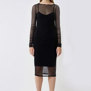 May include: A black midi dress with a sheer mesh overlay on the bodice and sleeves. The dress features a scoop neckline and spaghetti straps. The model is wearing black heeled sandals.