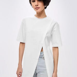 Puede incluir: Camiseta blanca de manga corta con diseño asimétrico. La camiseta tiene un frente cruzado con una abertura, combinada con vaqueros de lavado claro. La modelo tiene el pelo corto y oscuro.