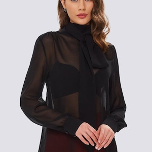 Może przedstawiać: Czarna, przezroczysta bluzka z wysokim dekoltem i długim krawatem. Top z długim rękawem ma mankiety zapinane na guziki i jest połączony z czerwonymi spodniami. Bluzka wykonana jest z lekkiej, przezroczystej tkaniny.