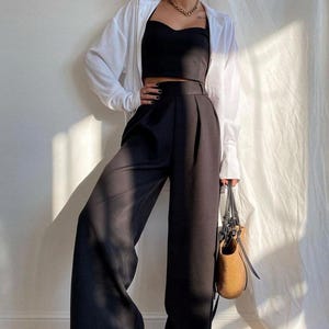Puede incluir: Una mujer lleva una camisa blanca abotonada sobre un top negro corto y pantalones negros de pierna ancha. Lleva un bolso tejido marrón con correas negras.