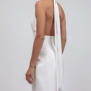 May include: A white halter jumpsuit with a flowing, open back design. The jumpsuit features a high neckline that ties at the back of the neck, with long, flowing fabric. The garment is made of a smooth, possibly satin-like material.