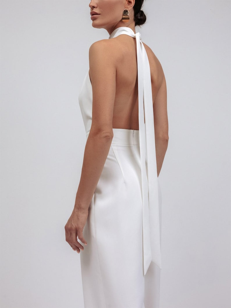 May include: A white halter jumpsuit with a flowing, open back design. The jumpsuit features a high neckline that ties at the back of the neck, with long, flowing fabric. The garment is made of a smooth, possibly satin-like material.