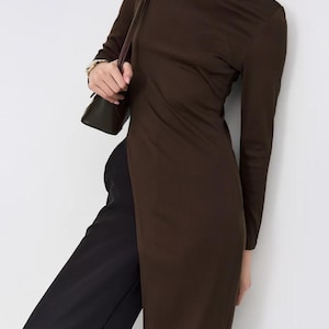 May include: A dark brown long-sleeved tunic dress featuring a high crew neckline and a long side slit that extends from the waist to the hem, paired with black trousers and a dark brown shoulder bag with a thin strap.