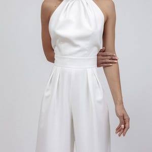 May include: A white halter-neck jumpsuit with a high neckline and wide-leg pants. The jumpsuit features a fitted waist and pleats in the pants. The fabric appears smooth and has a slight sheen. The overall style is elegant and minimalist.