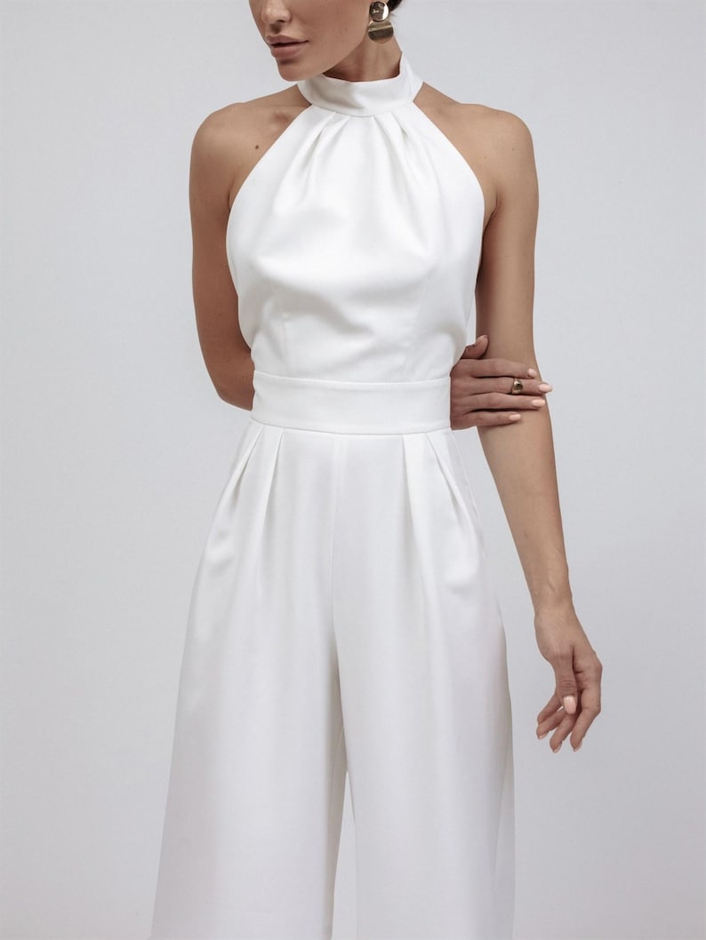 May include: A white halter-neck jumpsuit with a high neckline and wide-leg pants. The jumpsuit features a fitted waist and pleats in the pants. The fabric appears smooth and has a slight sheen. The overall style is elegant and minimalist.