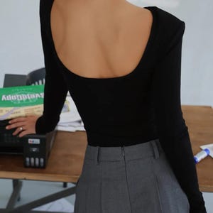 May include: A black long-sleeve top with a scoop back and padded shoulders. The top is paired with gray high-waisted trousers. The image is taken from behind, showcasing the back of the top and the trousers.