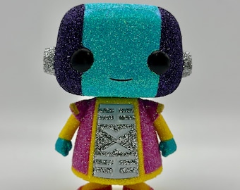 May include: A glittery Funko Pop figure of Zeno from Dragon Ball Super. The figure has a teal and purple head, black eyes, and a smiling expression. It wears a pink and yellow outfit with silver accents and teal gloves and shoes.