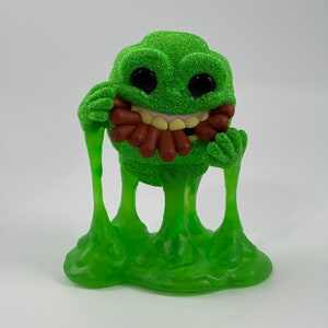 May include: Green and brown figurine of a monster with a toothy grin, holding a bunch of hot dogs in its mouth. The monster has gooey green legs and is standing on a green slime base.