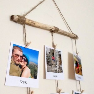 May include: A rustic wooden dowel rod with twine strung across it, holding up a collection of Polaroid-style photos. The photos are of various locations, including Crete, Buttermere, and a beach. Each photo has a handwritten label indicating the location.