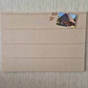 May include: A rectangular beige fabric-covered display board featuring four horizontal grooves. The board includes several small wooden clips and a single rectangular photograph depicting a traditional log cabin structure with a dark shingled roof.