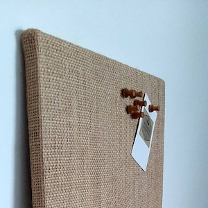 Op de afbeelding: Een bruin jute prikbord met houten duimspijkers en een wit kaartje met de tekst "The Everyday Essentials" in zwarte letters.