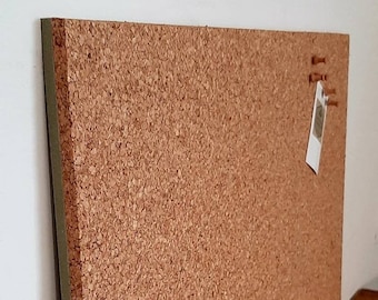 May include: A corkboard with a green border. The corkboard has a light brown color and is covered in small cork granules. There are three push pins and a small white tag attached to the corkboard.