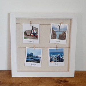 May include: A white framed photo display with four small travel photos clipped to twine. The photos depict scenic locations, including Madeira, Crete, and Scotland. The frame is set against a neutral background and a wooden surface.