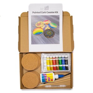 May include: Open cardboard box containing a Painted Cork Coaster Kit. The kit includes cork coasters, paint tubes, glue, brushes, and a booklet with painted coaster examples. The booklet shows coasters with rainbow, floral, and geometric designs.