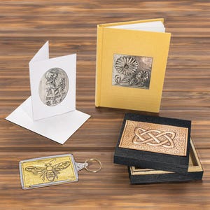 May include: A selection of handcrafted items: a yellow journal with a floral metal accent, a greeting card with an embossed figure, a gold bee keychain, and a small box with an embossed knot design.