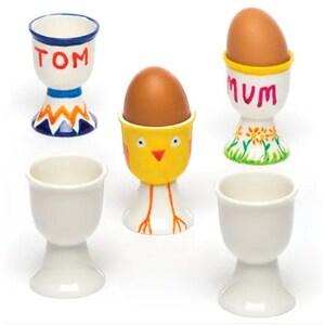 Könnte beinhalten: Eine Sammlung von Eierbechern aus Keramik, einige mit braunen Eiern. Ein Becher ist weiß mit dem Namen "TOM" in Rot, ein anderer ist gelb mit einem Küken-Design, und ein weiterer ist weiß mit "MUM" in Rot. Zwei schlichte weiße Eierbecher sind ebenfalls vorhanden.