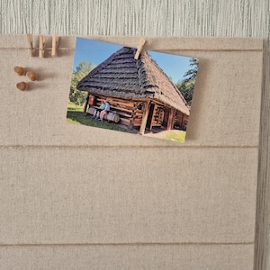 May include: A rectangular beige linen-covered display board featuring horizontal twine lines. A rectangular photograph of a rustic log cabin with a thatched roof is attached to the top twine with a small wooden clothespin, alongside three wooden pins.
