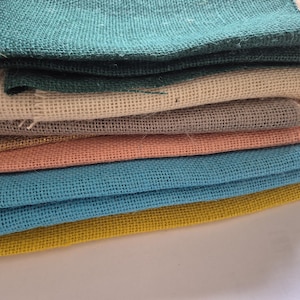 May include: A pile of colourful burlap fabric pieces in shades of teal, beige, grey, peach, blue, and yellow. The coarse texture of the fabric is visible, with each piece neatly folded and layered on top of each other.