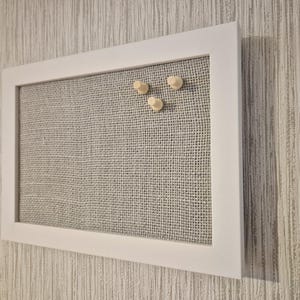 Op de afbeelding: Een wit omrand prikbord met een grijs jute oppervlak. Drie houten punaises zijn aan het bord bevestigd. Het rechthoekige bord is aan een gestructureerde muur bevestigd.