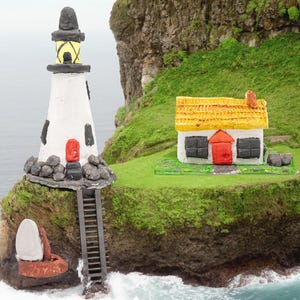 Könnte beinhalten: Eine Miniatur-Küstenlandschaft mit einem weißen Leuchtturm, schwarzem Dach und roter Tür, neben einem Cottage mit gelbem Strohdach. Eine Boje und eine Leiter sind ebenfalls vorhanden. Die Szene spielt auf einer grünen Klippe, mit dem Meer im Hintergrund.