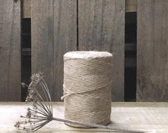 May include: A spool of natural twine sits on a wooden surface, with a dried flower in the foreground. The twine is tightly wound, and the texture is visible. The background is a weathered wooden panel.