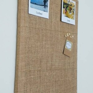 May include: A brown burlap cork board with two photos pinned to it. The photos are of a castle in Scotland and a building in Crete. The cork board has a small brown tag pinned to it with the text "Handmade with Love by WOODENLY".