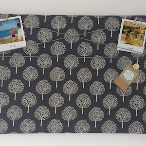 May include: A dark gray fabric bulletin board with a white tree pattern. The board has two photos pinned to it with clothespins. One photo shows a woman standing on a beach and the other shows a building with a stone facade. A brown tag with white text is pinned to the board. The text on the tag reads "Handmade with Love".