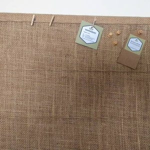 May include: A brown burlap bulletin board with two brown paper tags hanging from wooden clothespins. The tags have white text that reads "Rosemary and The Apothecary". There are also three wooden pushpins on the board.