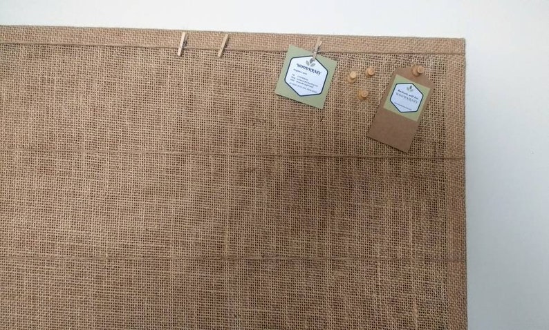 May include: A brown burlap bulletin board with two brown paper tags hanging from wooden clothespins. The tags have white text that reads "Rosemary and The Apothecary". There are also three wooden pushpins on the board.